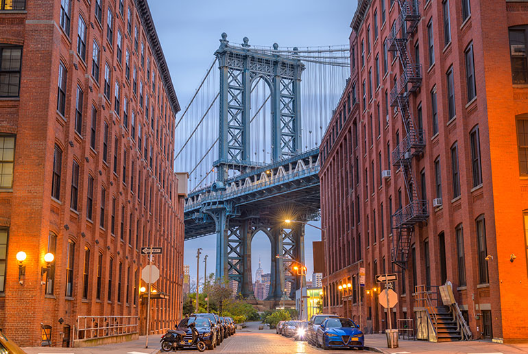 One of the blue towers of the Manhattan bridge as seen from downtown Brooklyn (dumbo).  One the left and right of the centered view industrial warehouses.  Through the whole in the bottom part of the tower a tiny empire state building.  